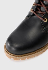 Stivale in pelle nera con lacci marroni e suola robusta di un leggero marrone, che mostra la parte frontale e parte del lato.