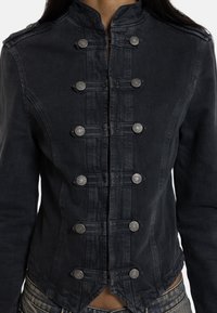 Κοντινό πλάνο ατόμου που φοράει εφαρμοστή σκούρα τζιν μπουφάν με διακοσμητικά ασημένια κουμπιά τακτοποιημένα σε διπλές κάθετες σειρές.