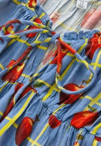 Modra bombažna obleka z rumenim mrežastim vzorcem, rdečimi potiski jabolk, nabrano teksturo in nastavljivimi trakovi na izrezu. Oznaka je vidna znotraj.