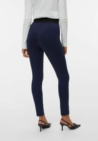 Marinblå leggings med en slät textur och hög midja, i kombination med svarta spetsiga klackar och en vit långärmad topp.