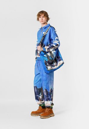 Sudadera azul con cremallera y diseño de montaña impreso, pantalones azules a juego y botas marrones. Incluye un bolso estampado con correas negras.