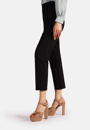 Sandali platform marroni con dettaglio annodato e cinturino alla caviglia, indossati con pantaloni neri aderenti. Texture liscia, con un design a tacco spesso.