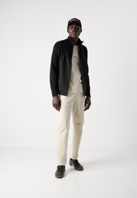 Jachetă neagră cu fermoar, tricou bej cu grafică, pantaloni bej deschis și teniși negri cu tălpi maronii. Căciulă neagră cu logo-ul "BOSS".