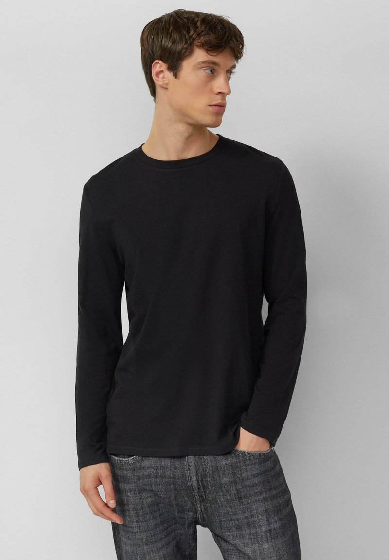 Czarny t-shirt z długim rękawem i okrągłym dekoltem, wykonany z miękkiego materiału, zestawiony z szarymi, teksturowanymi jeansami. Model stoi z rękami w kieszeniach.