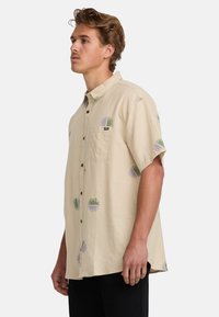 Camicia beige a maniche corte realizzata in tessuto leggero, con motivi circolari verdi e viola, chiusura frontale con bottoni e taschino sul petto.