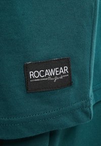 Tyrkysová bavlněná tkanina s žebrovanou strukturou; má černou obdélníkovou nášivku s nápisem "ROCAWEAR" a "ZALOŽENO New York 1999" v bílém písmu.