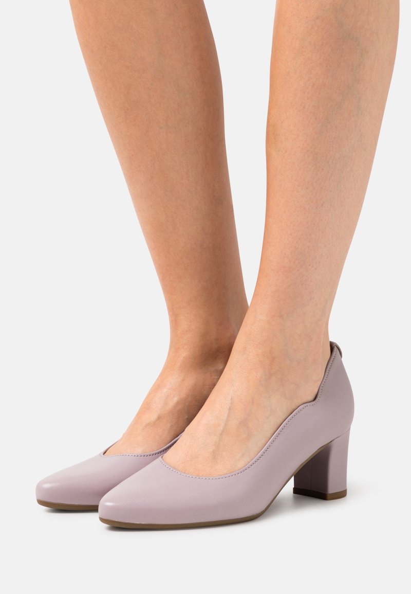 Jambes portant des chaussures fermées à talons moyens de couleur violet clair avec des bouts arrondis, sur un fond clair uni.