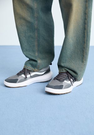 Gray athletic shoes with suede and mesh upper, white midsole, and rubber outsole. Paired with loose, faded blue denim pants.