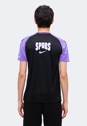 Person iført en sort og lilla sportstrøje med "SPURS" og et Nike-logo på ryggen, stående med fronten væk mod en ensfarvet baggrund.