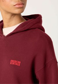 Bordeaux hoodie van zacht materiaal, met een voorzak en het logo "AMERICAN VINTAGE" in het rood op de borst, met een trekkoord bij de capuchon.