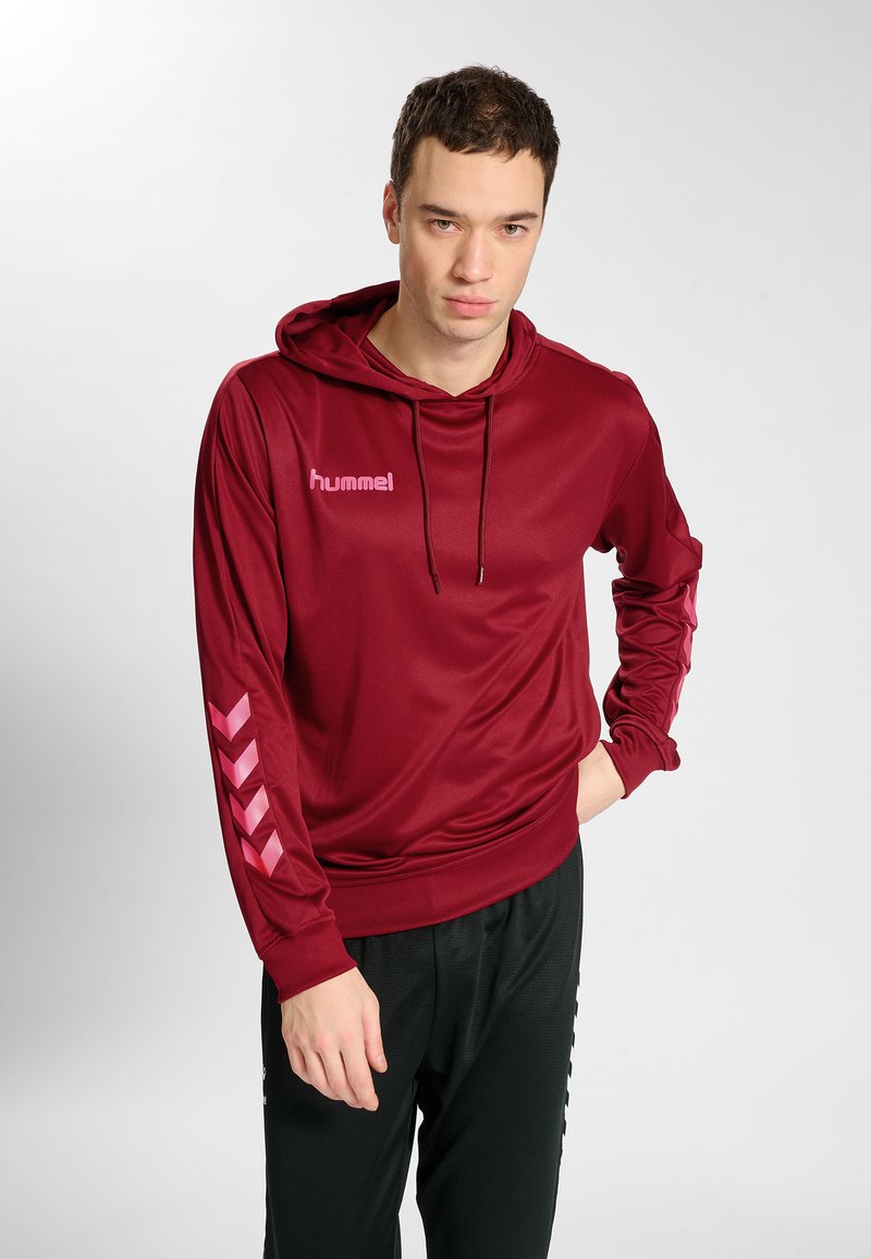 Bordowa sportowa bluza z gładką fakturą, z kapturem na sznurki, różowym logo i wzorem w jodełkę na rękawach. Całość dopełniają czarne spodnie.