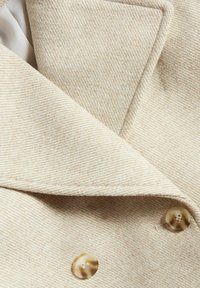 Vaaleanbeige kaksirivinen takki leveillä kauluksilla, valmistettu teksturoidusta kankaasta. Takissa on kaksi suurta vaaleanruskeaa nappia ja hienovarainen raidoitus.