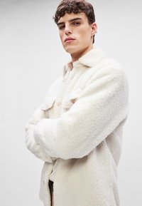 Kremfarget fleece skjortejakke med krage, knappelukking, to brystlommer og en teksturert, myk overflate.