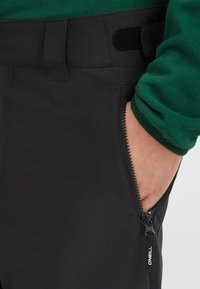 Svarta byxor med en jämn, matt yta. Har en sidkokficka och en kardborrebanddetalj vid midjan.