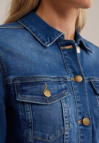 Jeansjacka i blått med spetsig krage, mässingsknappar och två bröstfickor. Har kontrasterande sömmar och en texturerad yta.