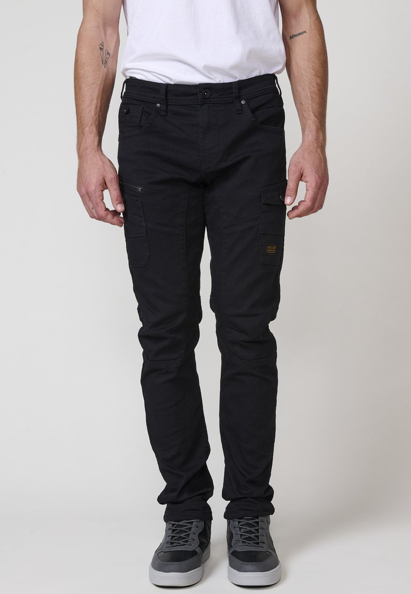 Svarta cargo-byxor i bomullsblandning med flera fickor, slim-fit design och kontrasterande gult logotypsmärke på sidan. Stylade med grå sneakers.