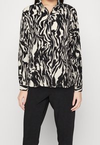 Blusa de manga larga en negro y crema con un patrón abstracto de olas. Presenta un cuello clásico y cierre de botones, hecha de una tela suave.
