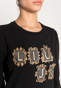 Sudadera negra con cuello redondo que presenta diseños ornamentados de letras en dorado y plateado, embellecidos con pedrería y patrones florales.