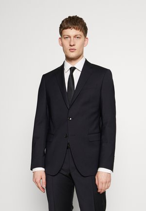Herrensakko in taillierter, marineblauer Farbe mit Reverskragen, einem Knopfverschluss, zwei vorderen Taschen und einer eleganten Silhouette. Weißes Hemd und schwarze Krawatte.