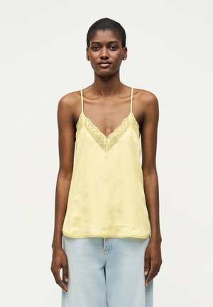 Kobieta w bladożółtej satynowej koszulce na ramiączkach z koronkową lamówką i jasnoniebieskich dżinsach, stojąca na jednolitym białym tle.