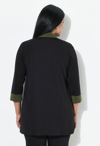 Zwarte tuniek met een groene kraag en manchetten. Kenmerkt zich door driekwartmouwen, een losvallende pasvorm en een gladde textuur. Geen zichtbare patronen of hardware.
