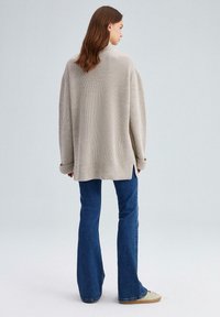 Lichtbeige oversized gebreide trui met een ribbelstructuur, featuring een hoge hals en zijsplitten, gecombineerd met uitlopende blauwe jeans.