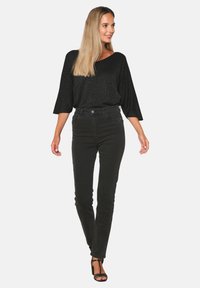 Čierny lesklý top s širokými rukávmi v kombinácii s čiernymi vysokými, priliehavými džínsami. Modelka má na sebe čierne remienkové podpätky, vlasy má upravené rovno.