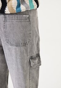 Grijze denim cargobroek met een patchzak aan de zijkant, voorzien van een merklabel. De stof vertoont een subtiele gewassen textuur.