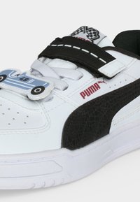 Λευκό και μαύρο αθλητικό παπούτσι Puma με λουράκι βέλκρο, σχέδιο αγωνιστικού αυτοκινήτου στα κορδόνια και λογότυπο σημαιούλας σκακιέρας στη γλώσσα.