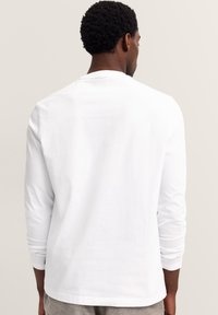 Mann mit kurzen lockigen Haaren, der ein schlichtes weißes Langarmshirt trägt und mit dem Rücken zu einem hellen neutralen Hintergrund steht.