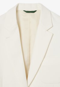 Blazer bianco su misura con revers a la francese, bottone singolo sul risvolto sinistro e taschino sul lato destro.