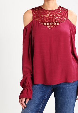 Kobieta ubrana w bordową bluzkę z wycięciami na ramionach ozdobioną koronką na górnej części piersi oraz długimi rękawami, w połączeniu z niebieskimi dżinsami.