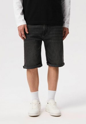 Mężczyzna ubrany w czarne podwinięte szorty jeansowe, białe trampki, białe skarpetki do kostek, czarny top i białą koszulę z długim rękawem, stojący na białym tle.