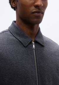 Σκούρο γκρι μπουφάν με φερμουάρ και γιακά, με λεία υφή υφάσματος και ασημί λεπτομέρεια φερμουάρ στο μπροστινό μέρος.