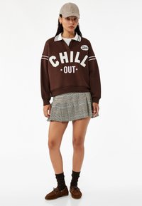 Brązowa bluza z napisem "CHILL OUT", biała szyja i paski. Szara kratkowana mini spódniczka, brązowe buty i ciemne skarpetki dopełniają strój.