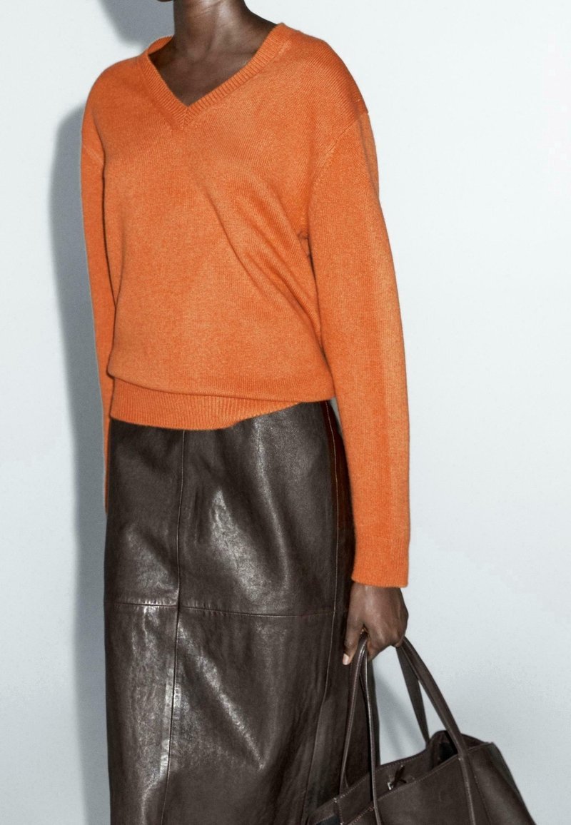 Personne portant un pull orange à col en V et une jupe en cuir marron foncé, tenant un sac cabas en cuir marron foncé sur un fond uni.