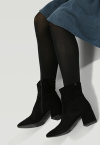 Černé semišové kotníkové boty s špičatou špičkou, robustním podpatkem a bočním zipem. Mají sklopený horní okraj a diskrétní logo značky.