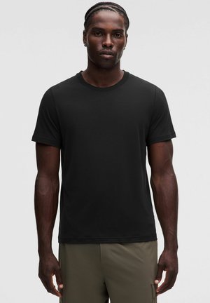 Hombre con el pelo trenzado que lleva una camiseta negra lisa de manga corta y pantalones verde oliva sobre un fondo gris claro.