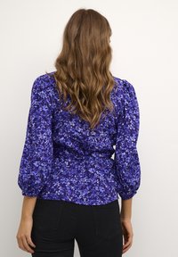 Blouse à imprimé floral bleu avec manches longues bouffantes, taille cintrée et tissu à texture douce. Le motif présente différentes nuances de bleu.