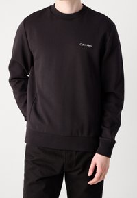 Sudadera negra hecha de tela suave, con cuello redondo, mangas largas y un pequeño logotipo blanco en el lado izquierdo del pecho.