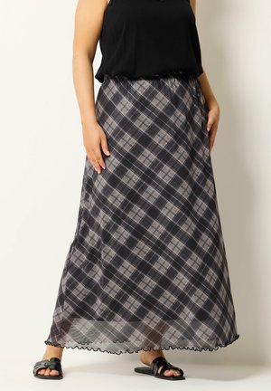 Kvinde iført sort ærmeløs top, lang sort- og gråternet nederdel og sorte slidssandaler, stående mod en ensfarvet baggrund.