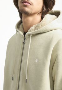 Sudadera verde clara con cierre de cremallera, forro de crema suave y un pequeño logo en el pecho. Tejido suave con un ajuste relajado.