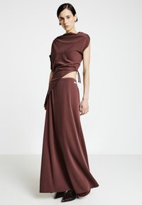 Robe longue bordeaux avec un haut drapé et une coupe asymétrique, dotée de liens à la taille et d'une texture lisse. Mannequin portant des talons pointus.