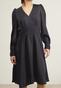 Svart klänning med långa, puffiga ärmar, V-ringning och en figursydd bodice. Flödande kjol som når mitt på låret, med strukturerad tyg.