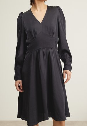 Schwarzes Kleid mit langen, puffigen Ärmeln, V-Ausschnitt und tailliertem Oberteil. Fließender Rock, der bis zur Mitte des Oberschenkels reicht, aus strukturiertem Stoff.