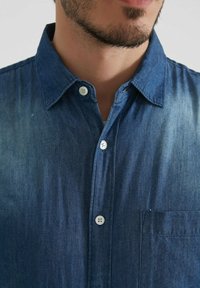 Chemise en denim bleu foncé avec une texture délavée, dotée d'un col à boutons, d'une poche avant et de boutons blancs.