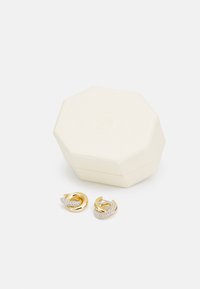 Orecchini tono oro e argento con accenti scintillanti, esposti accanto a una scatola per gioielli cremosa di forma ottagonale. La scatola ha una superficie texturizzata.