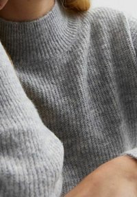 Gros plan d'une personne portant un pull en maille gris doux et côtelé, avec un col montant et des manches longues.