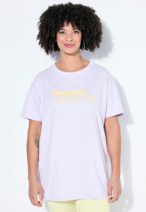 Donna sorridente con capelli ricci che indossa orecchini a cerchio, una maglietta viola chiaro con la scritta "felice è il nuovo ricco" e pantaloni giallo chiaro, in piedi davanti a uno sfondo bianco.