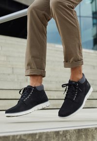 Black suede ankle boots with gray accents, a white sole, and lace-up design. Featured with rolled-up khaki pants, displayed against stairs.